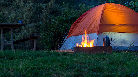 A fire in a designated fire pit with a Blue Coffee pot being heated. A tent with light glow and part of a picnic table with coffee cup fill out the picture, with bushes in the background. This is at a State Park in Oregon.