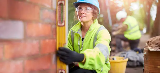 a young female bricklayer building a wall and using her level to check it .