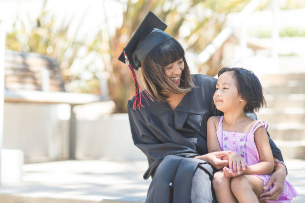 Graduation Day! Attractive Filipino woman in graduation robe and cap sits on steps with her young daughter after graduation. They are looking at each other affectionately and holding hands. nontraditional student photos stock pictures, royalty-free photos & images