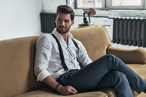 Handsome young man in white shirt and suspenders looking away and smiling while sitting on sofa