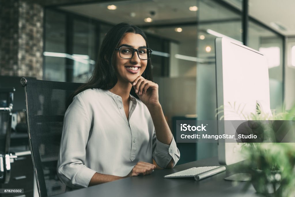 Cheerful young businesswoman working in office Portrait of cheerful young Latin-American businesswoman wearing glasses sitting at her workplace with computer in office looking at camera with hand on chin and smiling Indian Ethnicity Stock Photo
