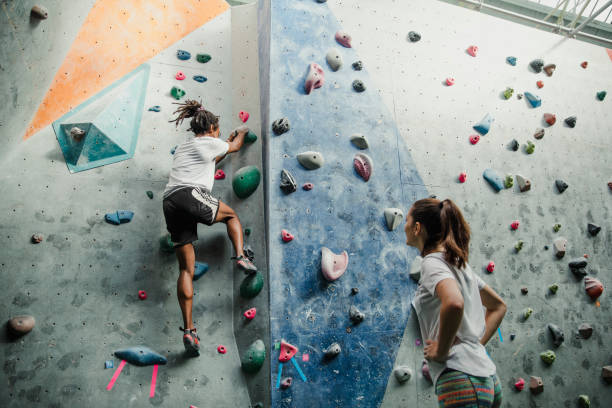 Intense Climbing Session Together Two people are enjoying an intense climbing session at an indoor rock climbing centre. The man is climbing a wall and his friend is watching him with her hands on her hips. clambering stock pictures, royalty-free photos & images