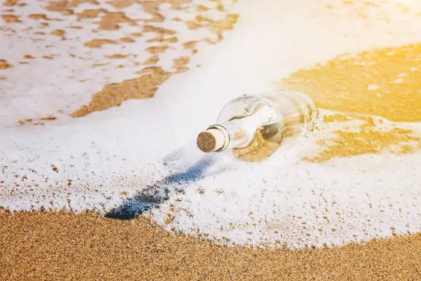 Message in a bottle washed up by the sea lying half submerged in the golden beach sand on the edge of the surf in a conceptual image of romance or a shipwreck with the glow of the sun in the corner