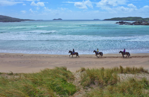 Loughshinny, Ireland, 1 May 2021 Skilled equestrians ride gracefully at a trot through shimmering waters, framed beautifully by vibrant clouds in the sky, creating a truly idyllic scene for all