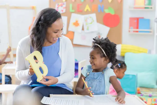 Cheerful mid adult female preschool teacher holds a letter "B" while working with a female student. The girl is also working on a worksheet.
