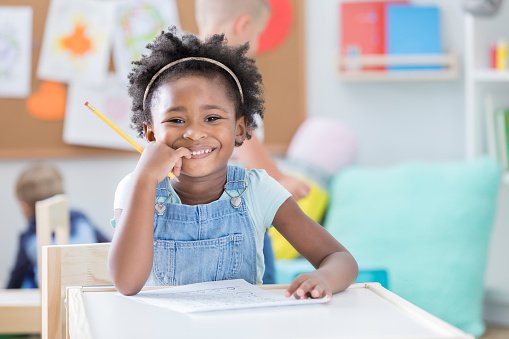 Adorable African American preschool student smiles confidently while working on a worksheet. She is holding a pencil and is smiling at the camera.