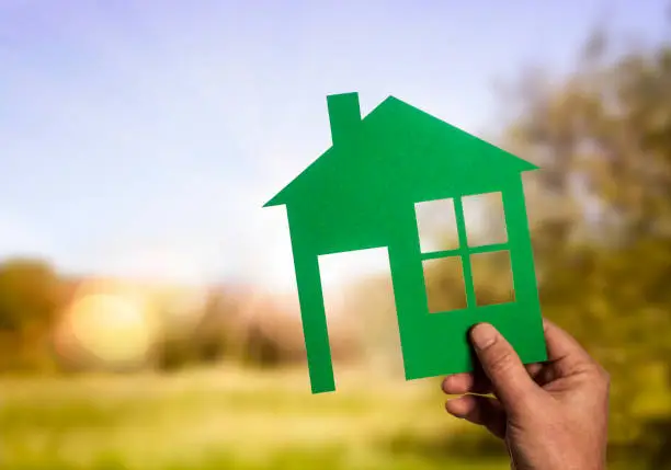 Hand with green silhouette of a house in front of a sunny landscape