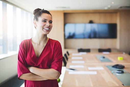 Smiling businesswoman standing by conference table. Confident young female executive is with arms crossed in board room. She is wearing red dress at office.