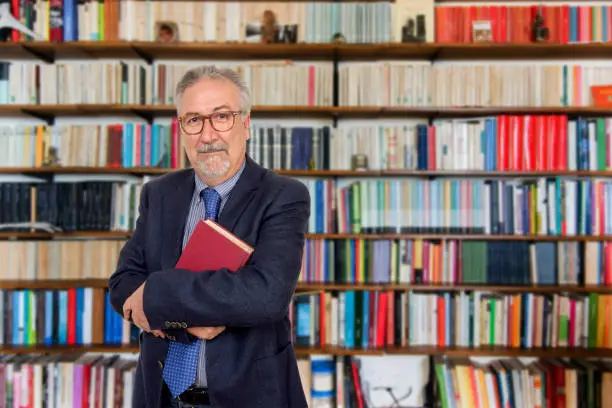 Photo of Senior teacher standing holding a book in front of a bookcase