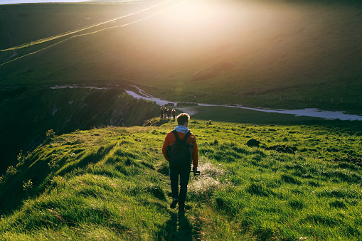 Young man in red jacket walking on a path towards Durdle Door alongside Jurassic Coast, Dorset, England during sunset. Green grass and hill soaked in the sun.