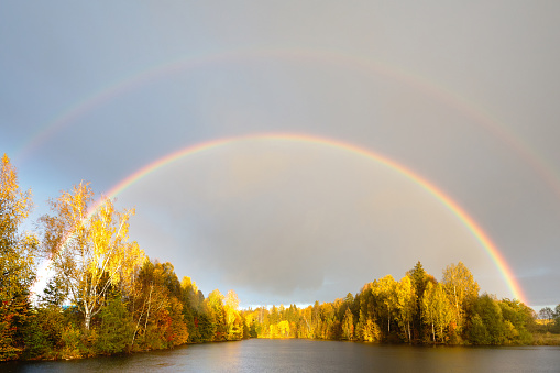 Rainbow over calm water of a forest lake. Beautiful clouds in the evening sky.