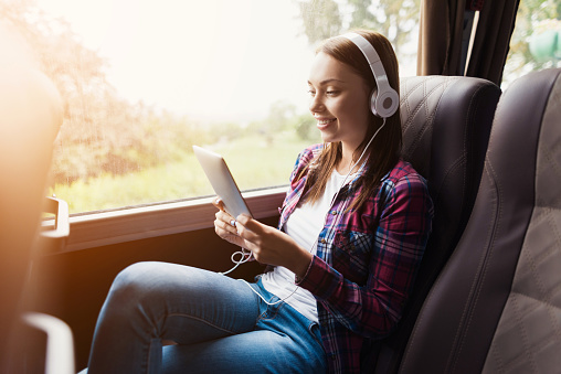 The woman on the passenger seat of the bus listens to music and looks at the tablet. She looks at the device's screen and smiles. Outside the window is a beautiful green landscape.