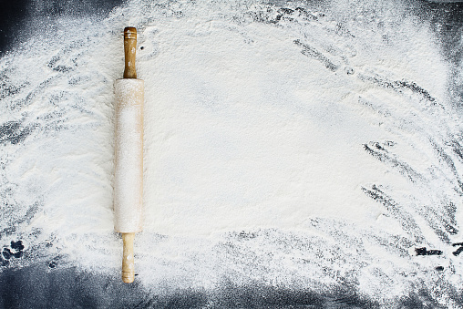 Old wooden rolling pin dusted with white flour over a flour covered dark background. Image shot from above.