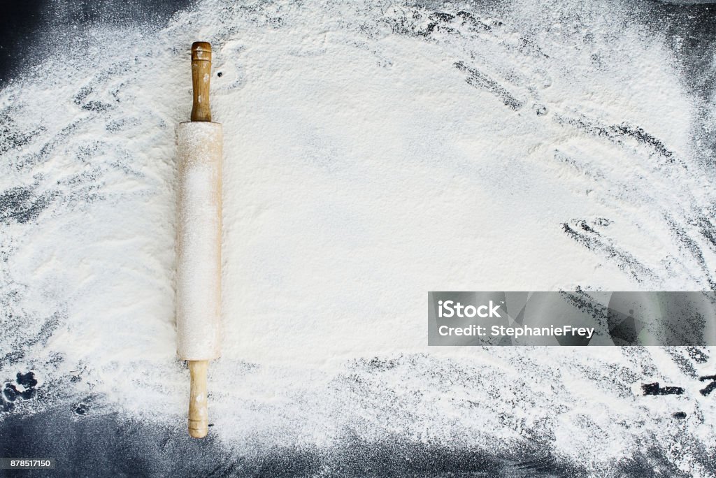 Rolling Pin Over Flour Background Old wooden rolling pin dusted with white flour over a flour covered dark background. Image shot from above. Flour Stock Photo