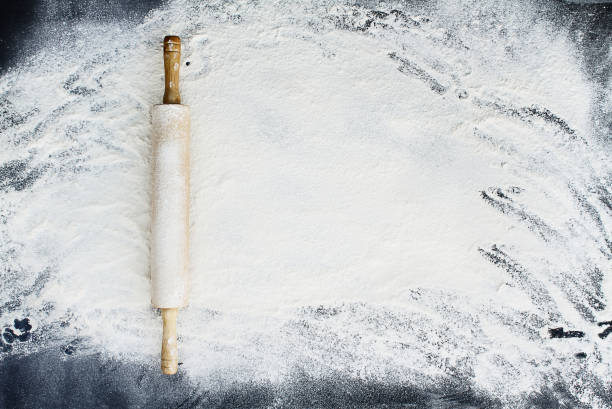 Rolling Pin Over Flour Background Old wooden rolling pin dusted with white flour over a flour covered dark background. Image shot from above. flour mess stock pictures, royalty-free photos & images