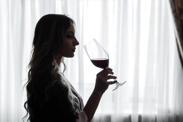 zmysłowa młoda kobieta ubrana w szatę pije czerwone wino z dużej kieliszka do wina, sylwetka w podświetleniu na tle okna - liquor store zdjęcia i obrazy z banku zdjęć