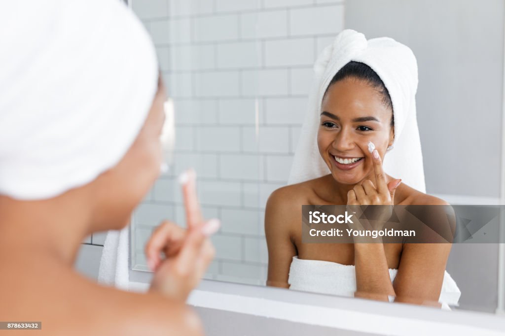 Portrait of a young smiling woman applying moisturiser to her face in the bathroom Moisturizer Stock Photo