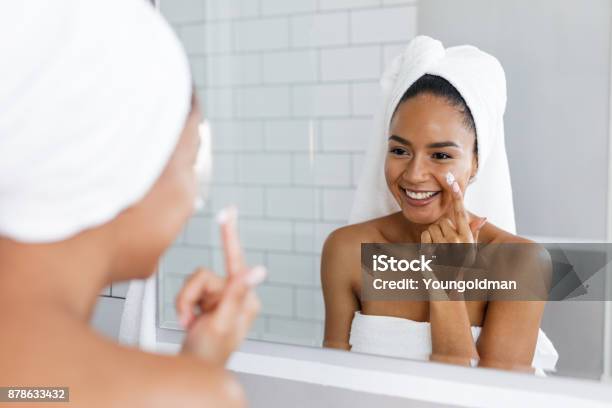 Portrait Of A Young Smiling Woman Applying Moisturiser To Her Face In The Bathroom Stock Photo - Download Image Now