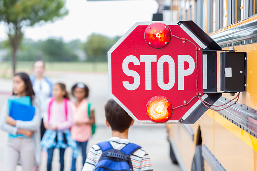 Close up of a flashing stop sign on a school bus. School children are blurred in the background. Focus is on the stop sign.