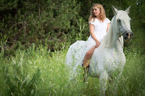 White Caucasian Blond Girl dressed in white sits on white horse in Field during the summer time.