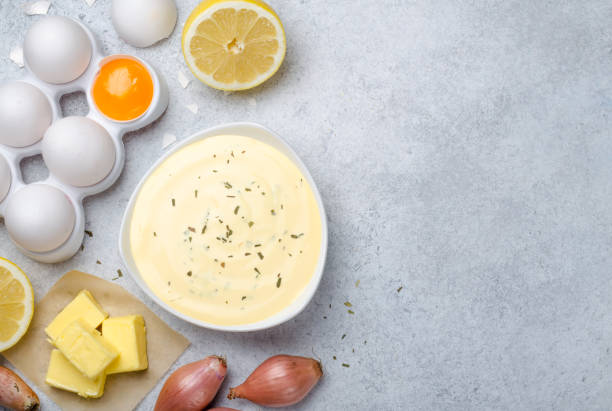 Basic french sauce bearnaise in a white bowl with ingredients, b Homemade basic french sauce bearnaise in a white bowl with ingredients, butter, shallot, lemon, eggs, on a light blue stone background with copy space, top view hollandaise sauce stock pictures, royalty-free photos & images