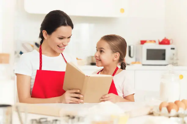 Mom and little daughter cook together in the kitchen at home. A woman and a girl in red aprons bake cookies and muffins. They have a happy family. They look at the recipes in the cookbook.