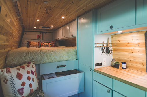 Living space of a young couple who live in a van. The sleeping area and bed are on the left. Underneath are cupboards and storage space. On the right are cabinets and a kitchen counter. The walls and ceilings are wood panels.