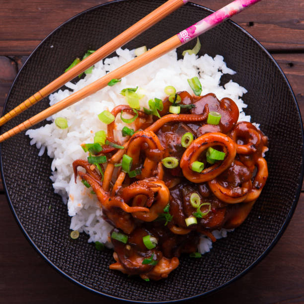 istock Spicy stir fried squid with rice and green onion and chopstick on black bowl on rustic brown wooden background 878803610