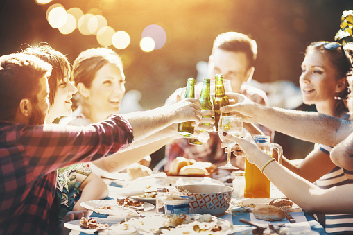 Closeup of group of young adults having a late afternoon garden party on a sunny summer day. They are having a feast and toasting with beers. There are three women and three men, some just partially visible, all released.