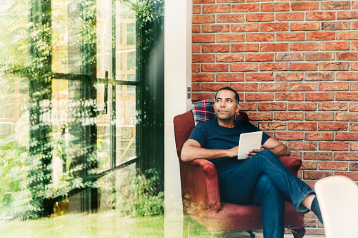 Thoughtful man sitting with digital tablet on armchair seen from window. Mature male is looking away against brick wall. He is relaxing at home.