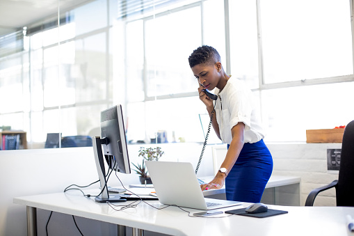 Mid adult businesswoman talking on landline phone while using laptop. Female professional is using technologies while standing in creative office. She is wearing smart casuals.