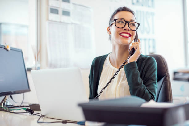 Businesswoman talking on landline phone in office Smiling young businesswoman talking on landline phone. Female professional is using telephone while looking away. She is working at desk in creative office. landline phone stock pictures, royalty-free photos & images