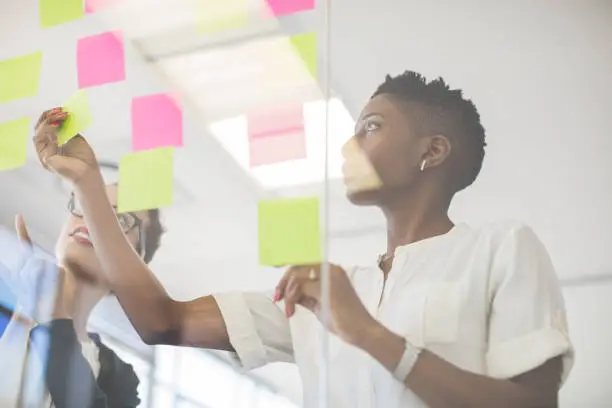 Businesswomen discussing over sticky notes on glass wall. Multi-ethnic female professionals are making strategy in creative office. They are wearing smart casuals.