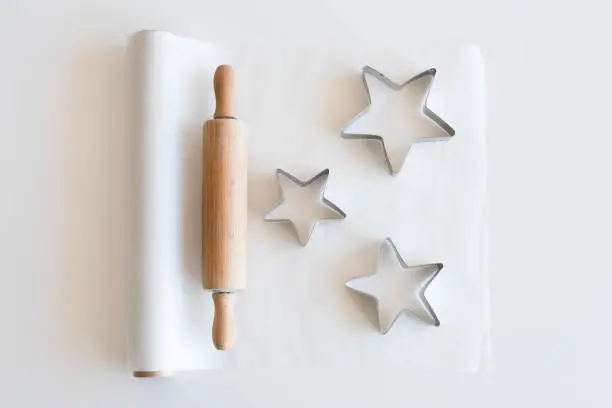 High angle view of baking paper, rolling pin and star shaped cookie cutters on white table - holiday baking concept