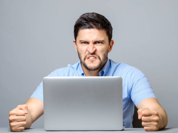 istock Angry man sitting at the table with laptop over gray background 878994406