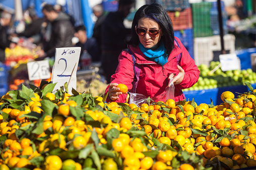 Adult woman wearing a red overcoat, sunglasses and a blue scarf shopping in farmer's market. She is choosing tangerines. She has black hair. Selective focus on model. Shot in daylight with a full frame DSLR camera.