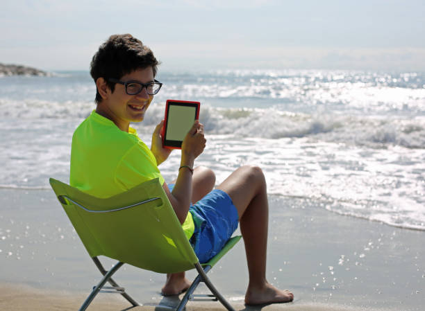 istock boy with glasses reads an ebook on the beach 879012450