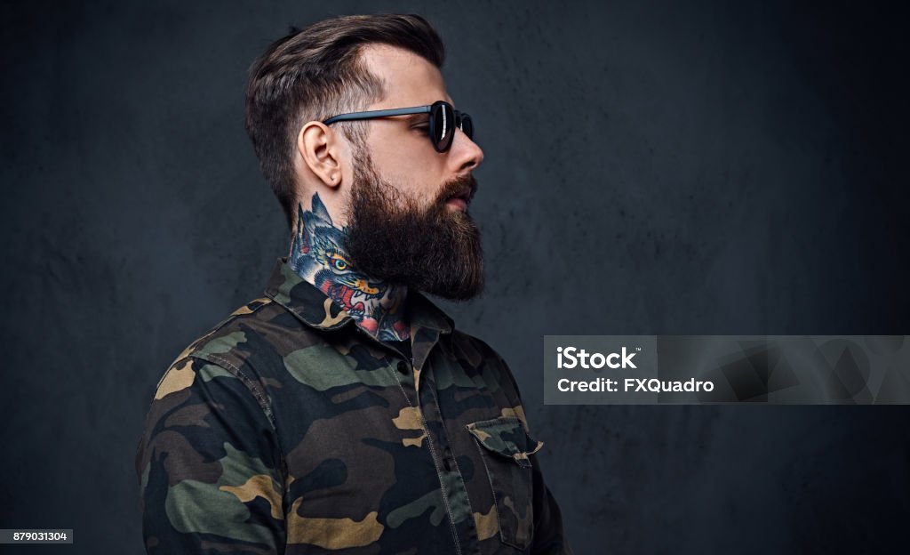 Portrait of bearded tattooed hipster male dressed in a military jacket. Portrait of bearded tattooed hipster male dressed in a military jacket over grey background. Barber Stock Photo