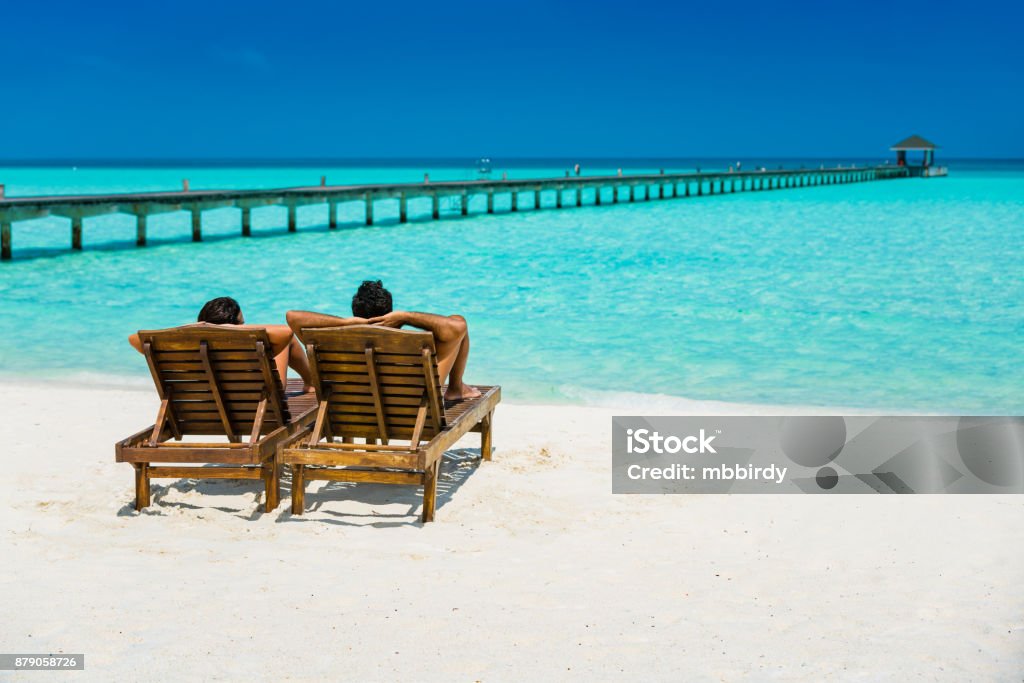 Couple enjoying holidays at Dhiffushi Holiday island, Maldives Couple enjoying holidays sitting on lounge chairs at tropical paradise beach at Dhiffushi Holiday island at South Ari atoll, Maldives. Beautiful turquoise Indian ocean sea with white sand. Luxury travel holidays background. Model and Property released. Beach Stock Photo