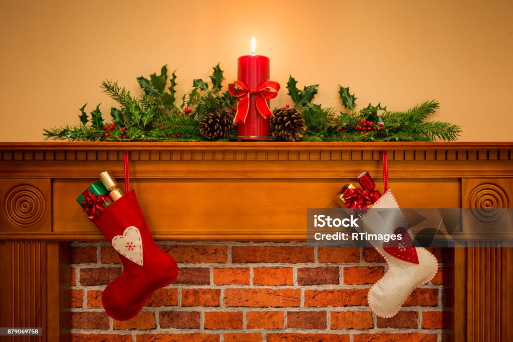 Christmas stockings hanging on the fireplace Two Christmas stockings with gift wrapped presents hanging on a mantelpiece over a fireplace, plus burning candle with festive garland including holly, fir and pine cones. Mantelpiece Stock Photo