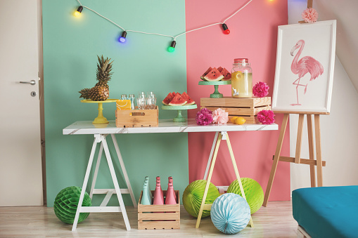 Juicy watermelon slices, pineapple, and fresh lemonade are served on summer party table. The wall behind the table is colored in green and pink, and decorated with colorful lights.