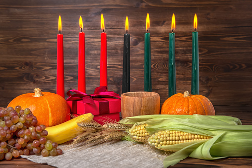 Kwanzaa festival concept with seven candles red, black and green, gift box, pumpkins, ears of wheat, grapes, corns, banana, bowl and fruits on wooden background, close up