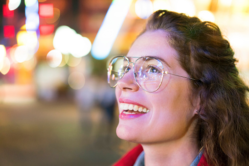 Close-up shot of a smiling young woman with eyeglasses amazed by flashing city lights.