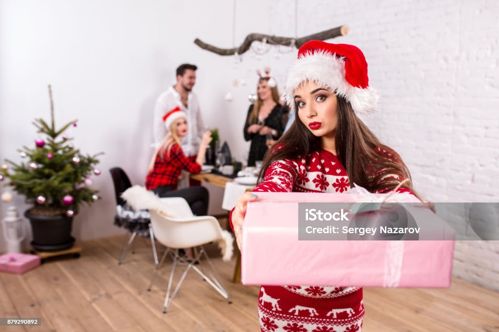 Portrait of a young woman with a pink giftbox at home on the foreground. Beautiful brunette in a Santa hat, red costume with deers Portrait of a young woman with a pink giftbox on the foreground. Beautiful brunette in a Santa hat, red costume with deers. Christmas party at home. Adult Stock Photo