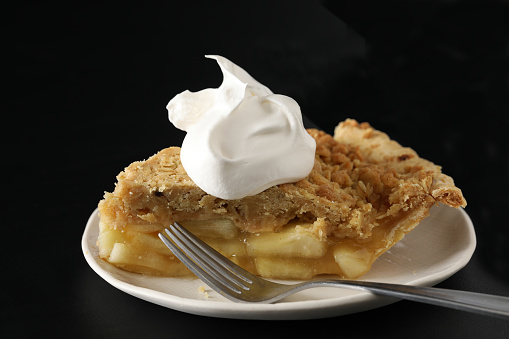 A close up horizontal photograph of an apple pie topped with whipped cream on a white plate with a fork.Isolated on black.