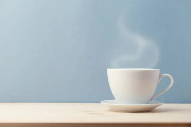 Small white cup on saucer with steam coming from hot delicious drink on blue background.