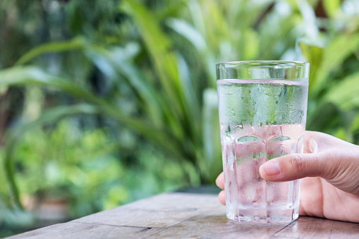 Closeup image of a hand holding a glass of cold water on wooden table with green nature background