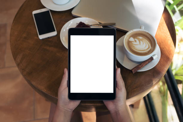 istock Top view mockup image of hands holding black tablet pc with blank white screen , smart phone , laptop , coffee cup and cake on wooden table in cafe 879527054