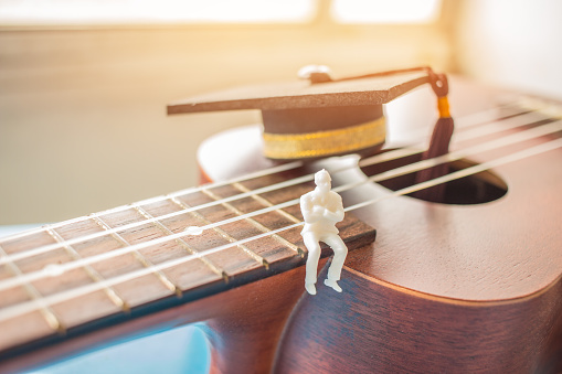 Miniature figurines Businessman sitting on ukulele with Graduation cap. Ukuleles is member lute family instruments with nylon stringed, played with bare thumb, fingertips. Concept of Music Education.