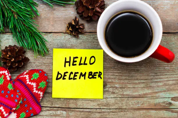 Photo of Hello December- handwriting in black ink on a sticky note with a cup of coffee and mittens, New Year resolutions concept